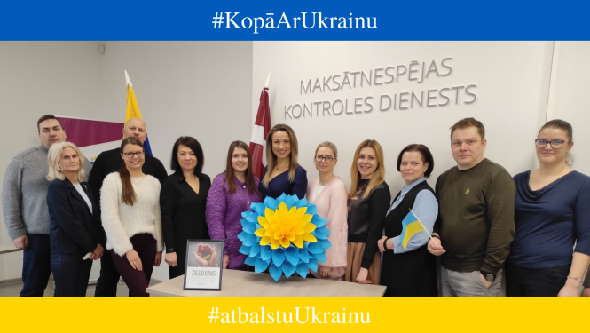 MKD kolektīvs stāv pie galda ar papīra puķi Ukrainas karoga krāsās. Aiz muguras Ukrainas un Latvijas karogi.