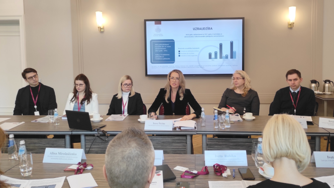 MKD direktore Inese Šteina pie galda, prezentācijas ekrāns aiz muguras