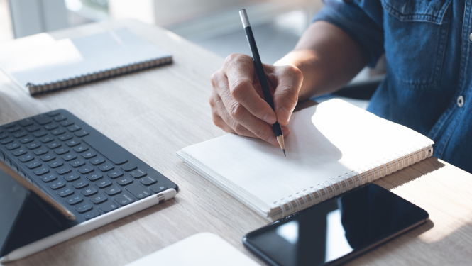 Cilvēks pie galda iepretim datoram raksta piezīmes